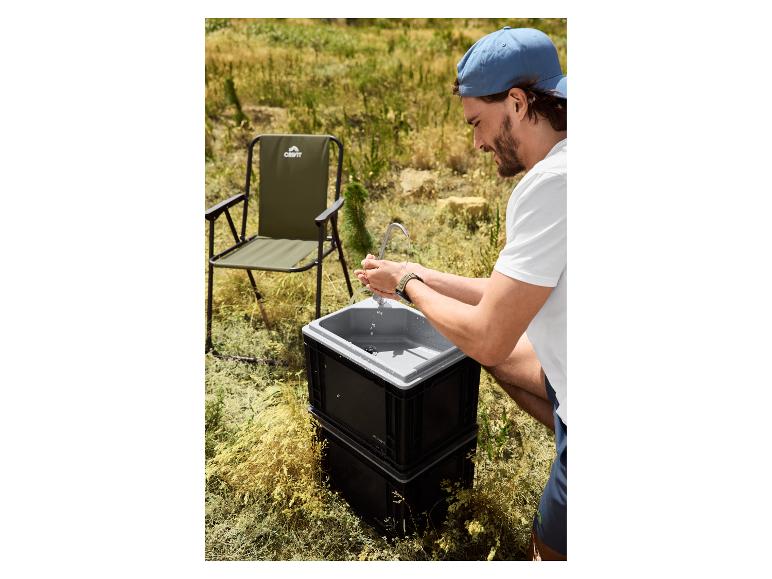 Mann wäscht Hände an einem tragbaren Waschbecken mit Campingstuhl im Hintergrund.