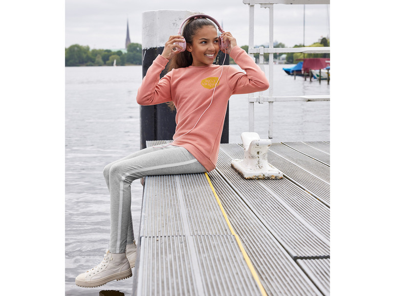 Ein Mädchen mit einem rosa Pullover, grauen Leggings und weißen Sneakern hört Musik auf einem Steg.