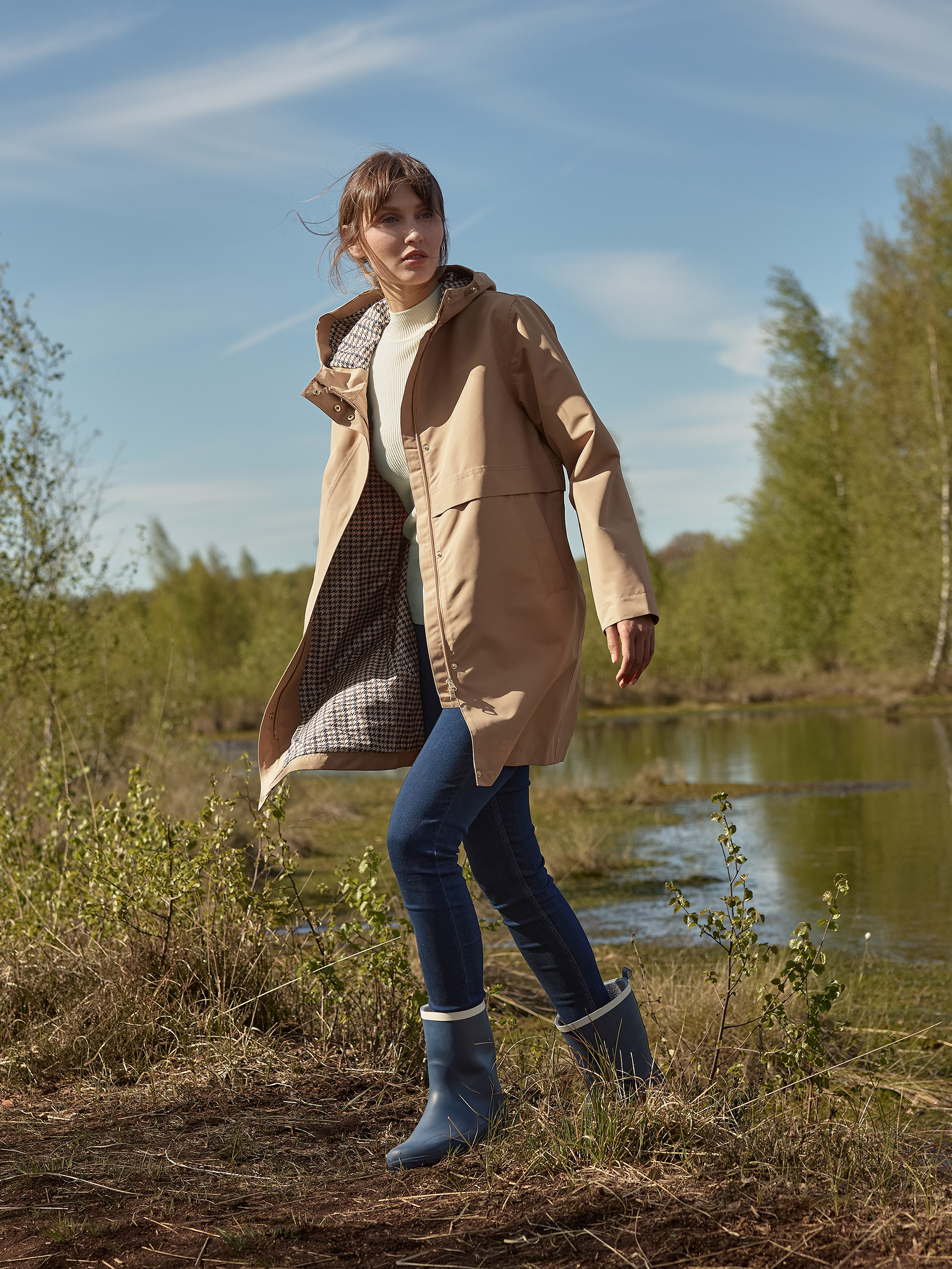 Frau in beigem Trenchcoat, Jeans und blauen Gummistiefeln, geht an einem See entlang.