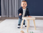 Ein Baby in einem blauen Pullover und Hose steht auf einem Holzhocker.