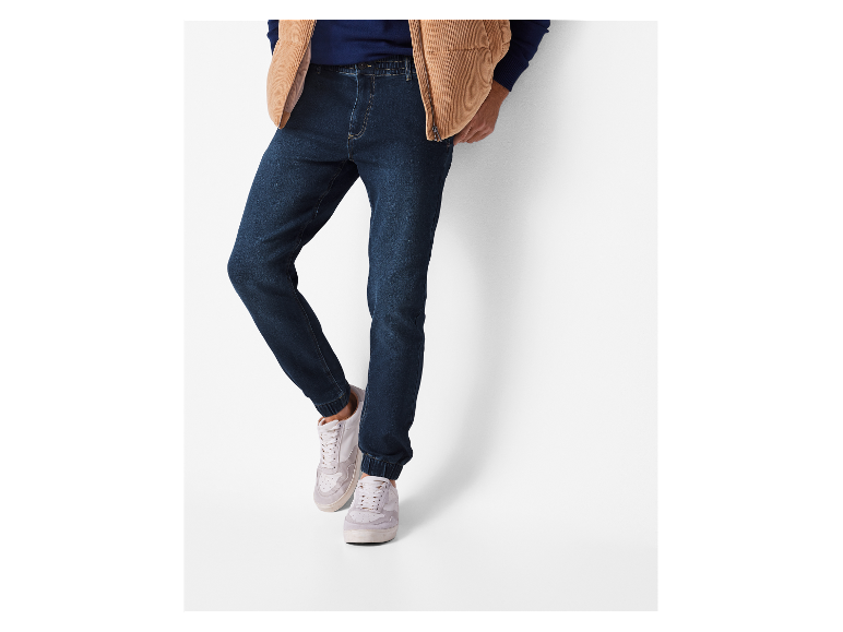 Mann trägt dunkelblaue Jeans, blauen Pullover und beige Steppweste.