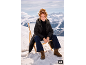 Frau in schwarzer Jacke und Jeans sitzt auf einem Stuhl in verschneiter Berglandschaft.