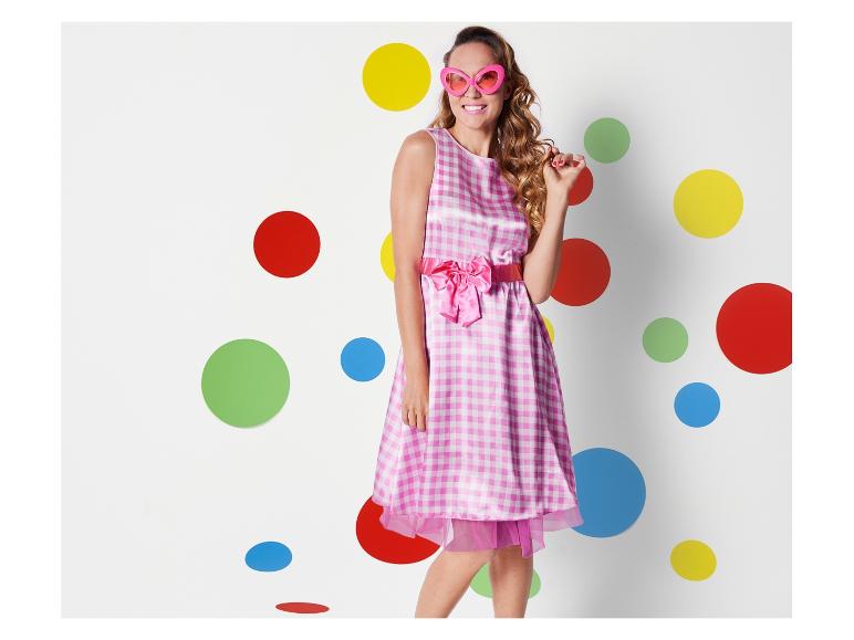 Eine Frau in einem rosafarbenen kariertes Kleid mit einer rosafarbenen Schleife steht vor einem Hintergrund mit bunten Punkten.
