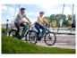 Ein lächelndes Paar fährt E-Bikes entlang einer sonnigen Promenade.