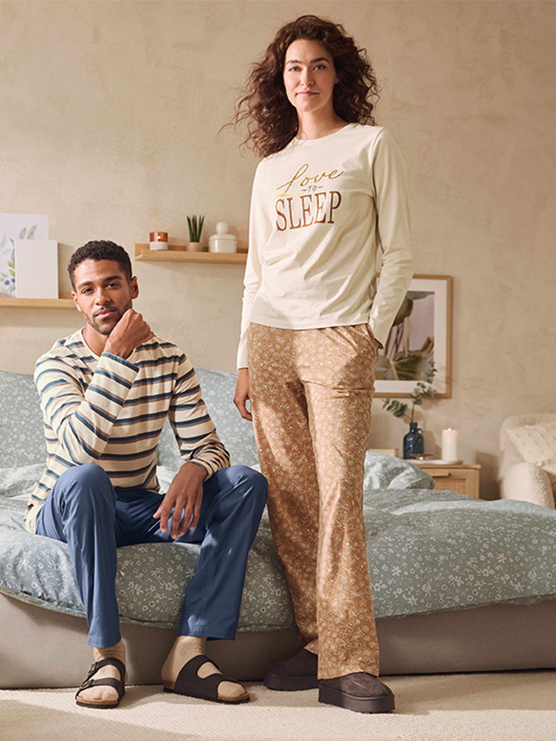 Ein Paar trägt passende Pyjamas, er in Streifen und blauen Hosen, sie in einem 'Love to Sleep' Oberteil und gemusterten Hosen.