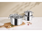 Set glänzender Edelstahl-Töpfe und -Pfannen auf einem Schneidebrett mit Knoblauch und Rosmarin.