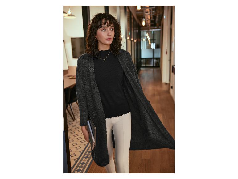Eine Frau trägt einen schwarzen Pullover, einen langen grauen Cardigan und weiße Hosen.