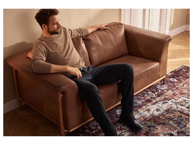 Ein Mann sitzt auf einem braunen Ledersofa, trägt einen beigen Pullover und schwarze Hose.