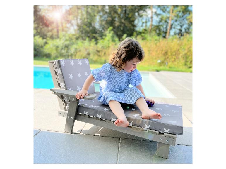 Kleinkind auf einem Holzliegestuhl mit grauen Kissen mit weißen Sternen neben einem Pool.