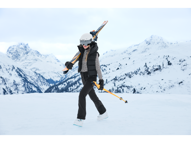 Eine Frau in stilvoller Skikleidung geht mit Skiern auf der Schulter durch verschneite Berge.
