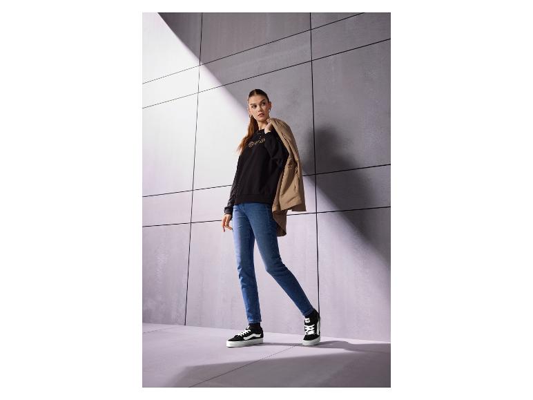 Frau in schwarzem bedrucktem Sweatshirt, Jeans und Turnschuhen, mit beiger Jacke über der Schulter.