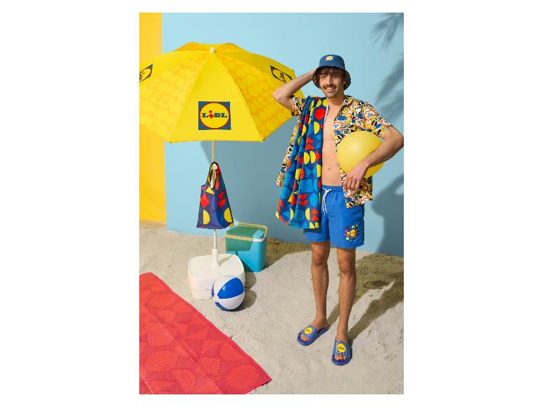 Mann in Sommerkleidung und Badeschlappen, mit Handtuch und Wasserball, unter einem Sonnenschirm.