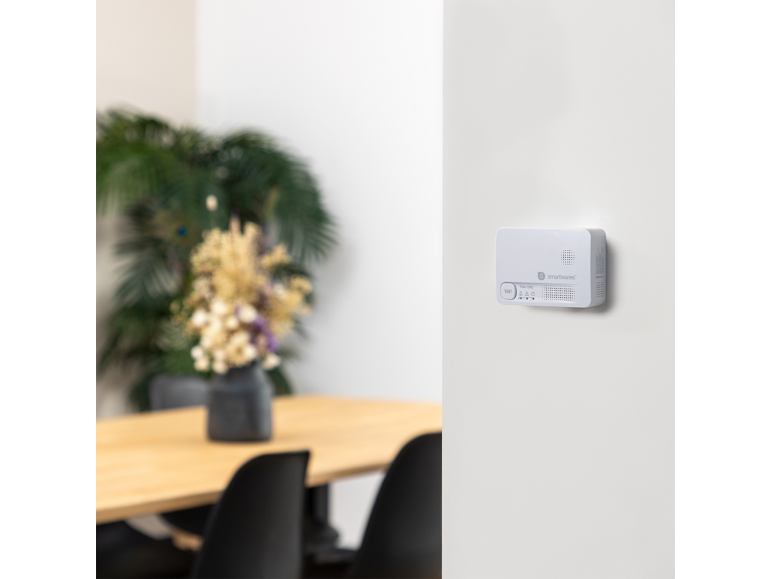 Weißer Rauchmelder an der Wand, mit Holztisch und Blumen im Hintergrund.