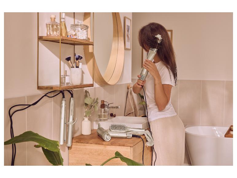 Frau stylt ihre Haare mit einer Warmluftbürste in einem modernen Badezimmer mit Haarstyling-Geräten und Kosmetik.