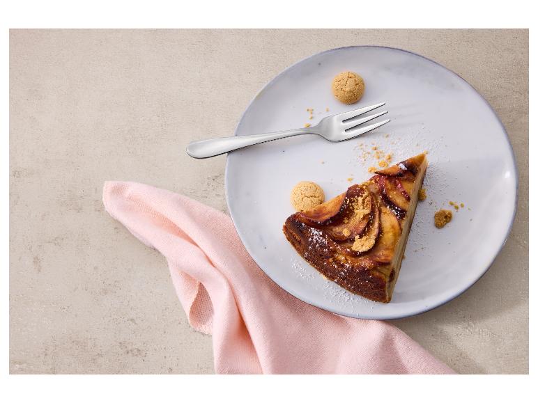 Ein Stück Apfelkuchen mit Gabel und Keksen auf einem Teller, vor einem rosa Tuch.