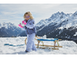 Mädchen in Skijacke, Hose und rosa Handschuhen, spielt mit Schnee neben einem Schlitten.