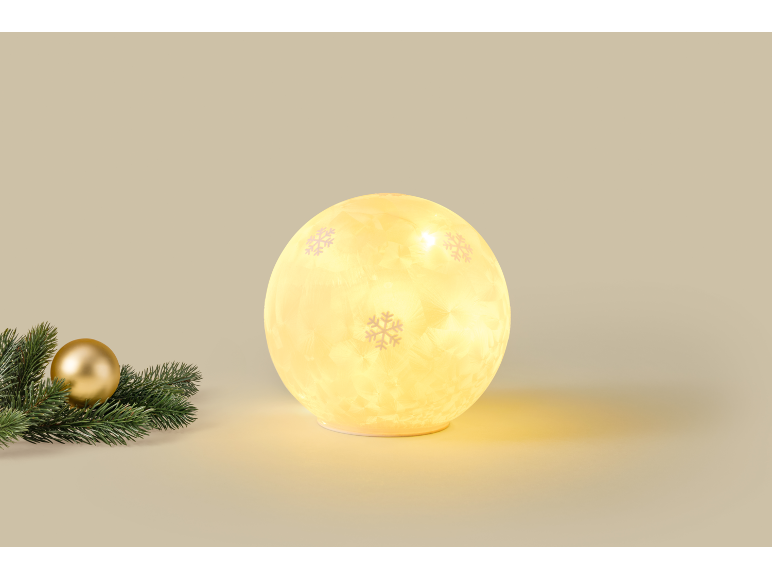Leuchtende kugelförmige Weihnachtslampe mit Schneeflockenmuster neben Tannenzweig und goldener Kugel