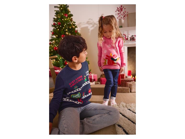 Zwei Kinder in Weihnachtspullovern, ein Junge auf dem Boden und ein Mädchen stehend, neben einem Weihnachtsbaum.