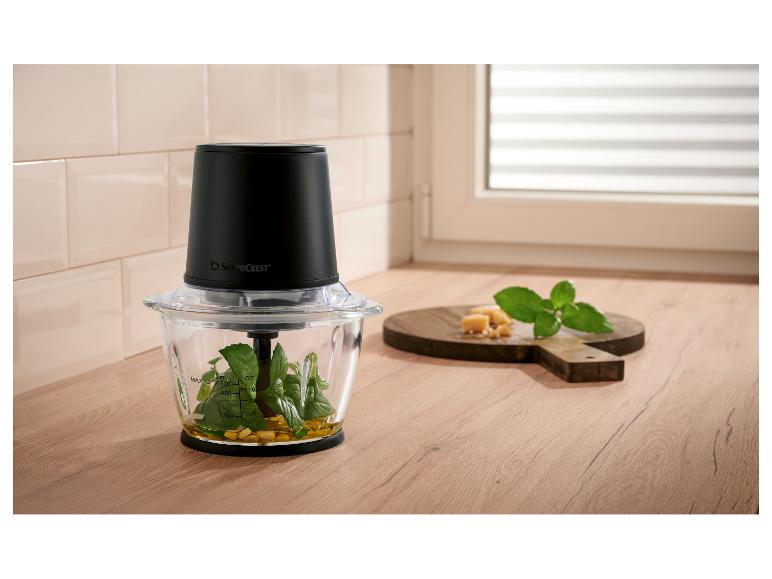 Zerkleinerer mit Basilikum, Öl und Nüssen, daneben Käse und Minze auf einem Holzbrett.