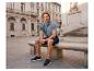 Mann in blauem Poloshirt und Shorts sitzt auf einer Steinbank auf einem Stadtplatz.