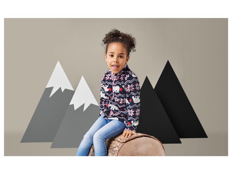 Ein Mädchen in einer blauen Fleecejacke mit Bärenmotiv und Jeans sitzt auf einem Baumstamm.