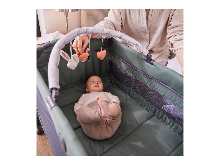 Baby liegt in Reisebett mit Spielzeug, Erwachsener hält eines der Spielzeuge.