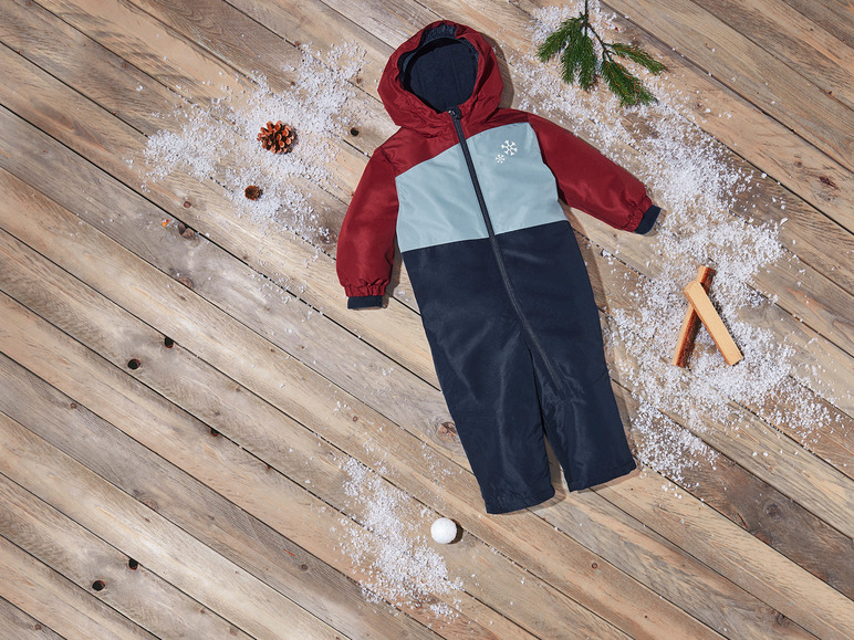 Ein rot-blau-grauer Winteroverall für Kinder auf einem Holzboden mit Schnee.