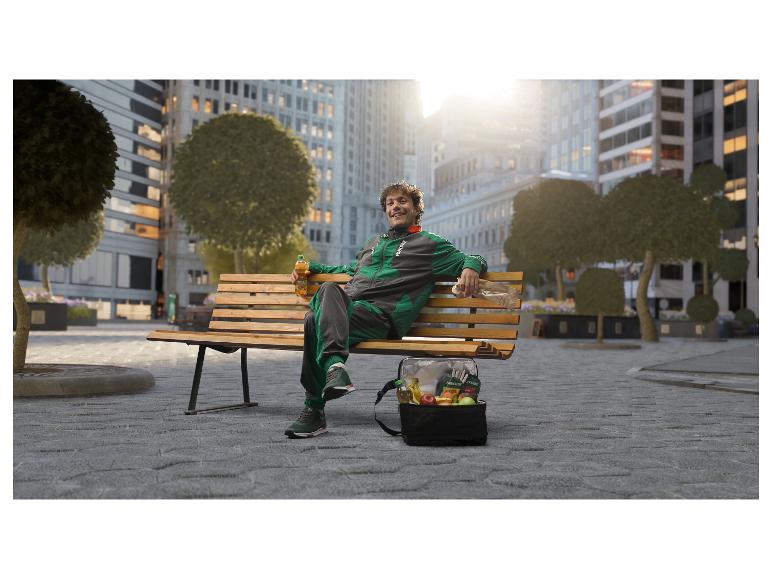 Mann im Trainingsanzug auf einer Parkbank mit einer Kühltasche voller frischer Früchte und einem Getränk.