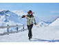 Frau in Skijacke, Skihose, Helm und Skibrille trägt Skier und Stöcke in verschneiter Berglandschaft.