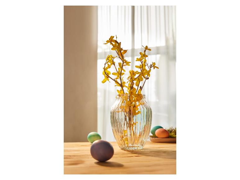 Gelbe Blumen in einer Glasvase mit Ostereiern auf einem Holztisch.