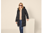 Frau in einer schwarzen langen Steppjacke, beigem Pullover und Jeans.