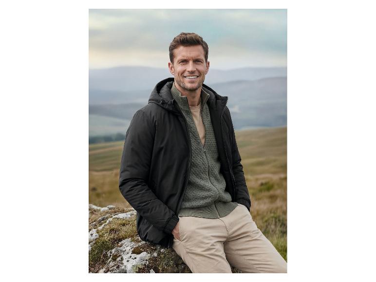 Mann in schwarzer Jacke und grünem Pullover sitzt auf einem Felsen in bergiger Landschaft.