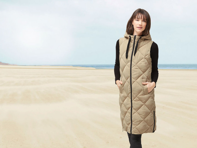 Eine Frau trägt eine lange, beige wattierte Weste mit Kapuze am Strand.