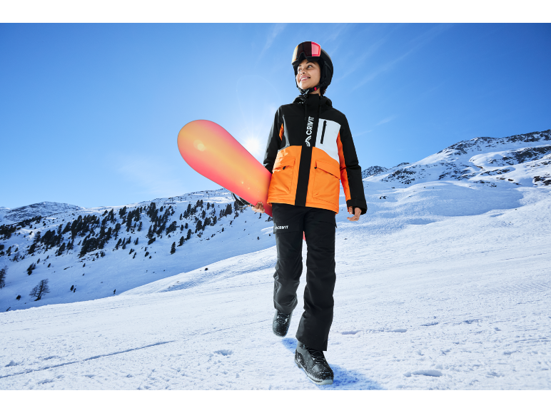 Junge Person in Skijacke und Hose mit Snowboard am verschneiten Berghang.