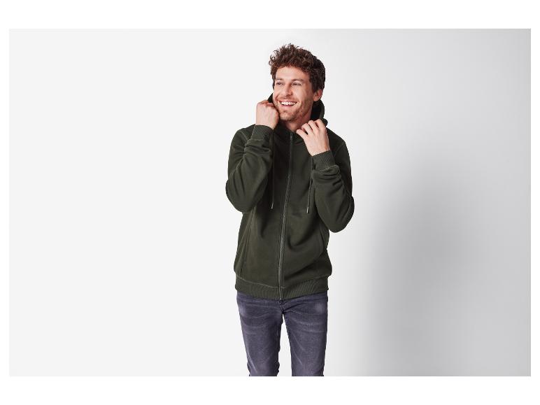 Lächelnder Mann in einem dunkelgrünen Kapuzenpullover mit Reißverschluss und grauen Jeans.