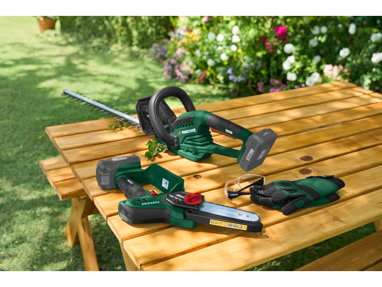 Akku-Heckenschere und Astsäge auf einem Holztisch im Garten mit Schutzbrille und Handschuhen.