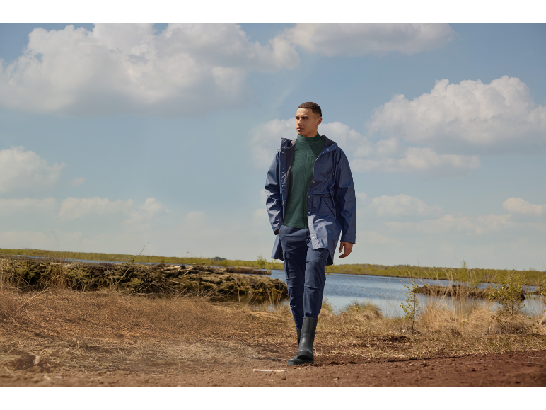 Mann in blauer Regenjacke und schwarzen Gummistiefeln in Naturlandschaft.