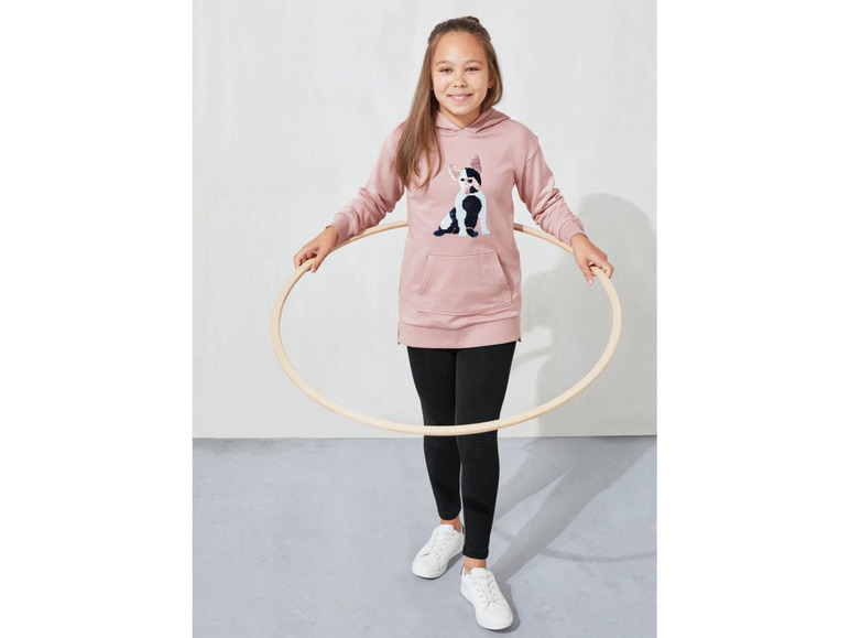 Ein Mädchen in einem rosa Hoodie mit Hundeaufdruck, schwarzen Leggings und weißen Sneakern hält einen Hula Hoop.