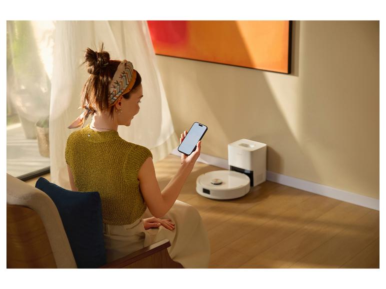 Frau steuert Saugroboter mit Smartphone in hellem Wohnzimmer.