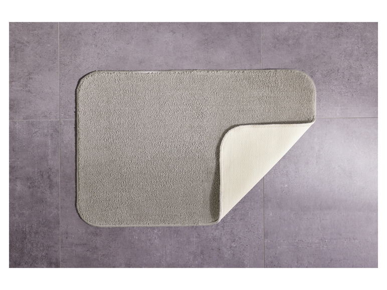 Ein grauer Badezimmerteppich mit beigefarbener Unterseite auf Fliesen.