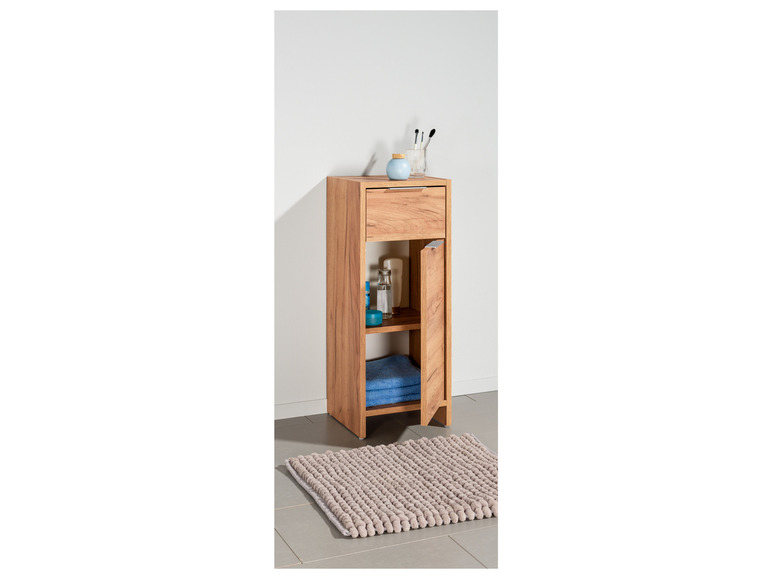 Ein Badezimmer-Schrank aus Holz mit einer Schublade, einem Regal und einer Tür, mit einem Badezimmerteppich darunter.