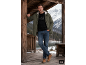 Mann in grüner Jacke, schwarzem Shirt und Jeans, lehnt an Holzsäule im Freien.