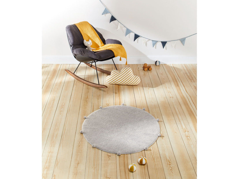 Ein runder grauer Teppich mit Pompons für Babys mit einem schwarzen Holz-Schaukelstuhl.