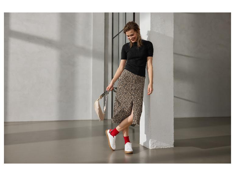 Frau in schwarzer Bluse, Leopardenrock, roten Socken und weißen Sneakern, hält eine Tasche.