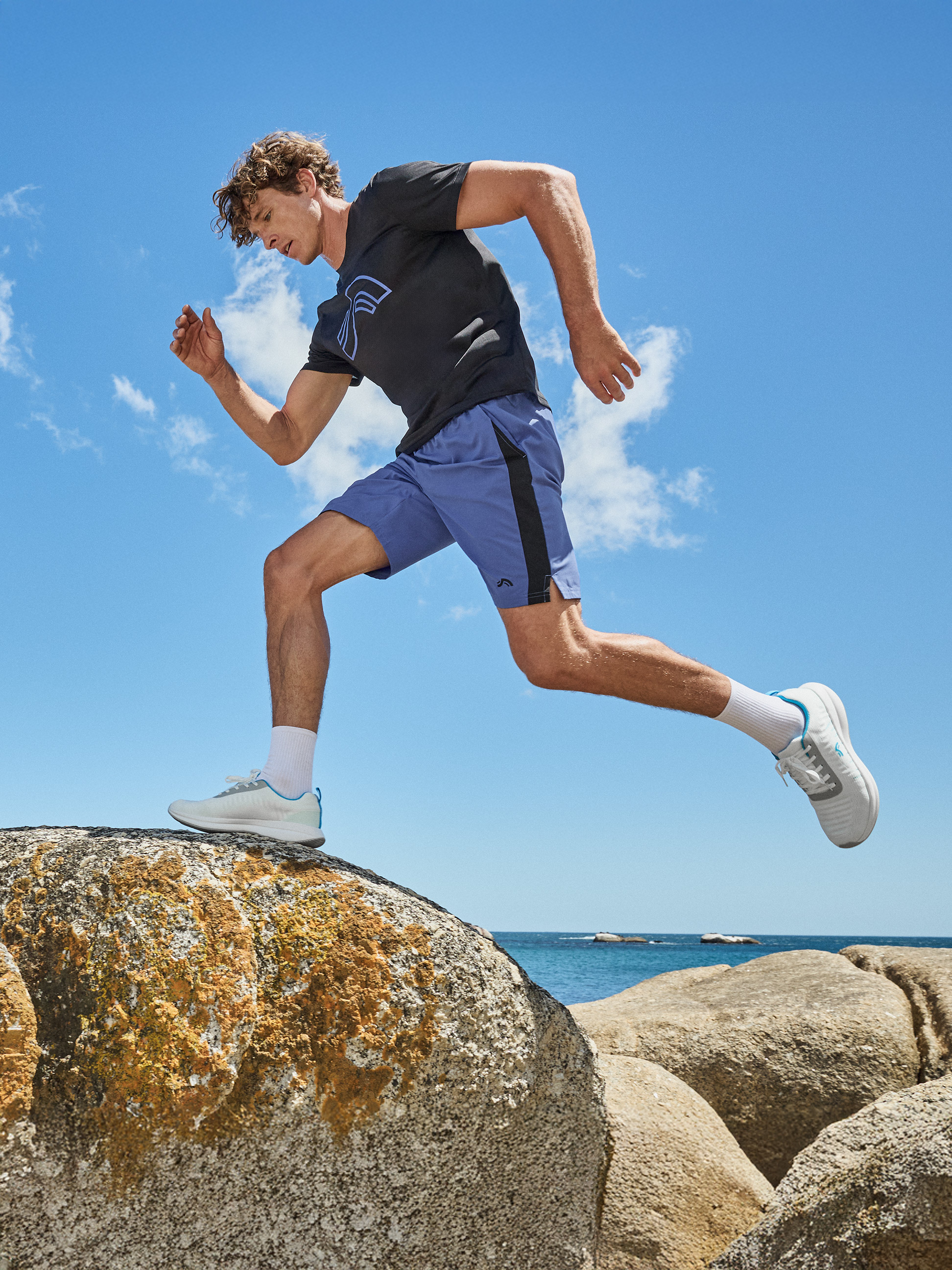 Mann in schwarzem T-Shirt und blauen Shorts läuft auf Felsen am Strand.