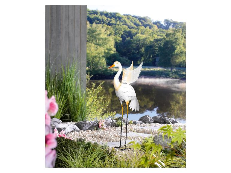 Eine weiße Metall-Reiher mit ausgebreiteten Flügeln in einer Gartenlandschaft.