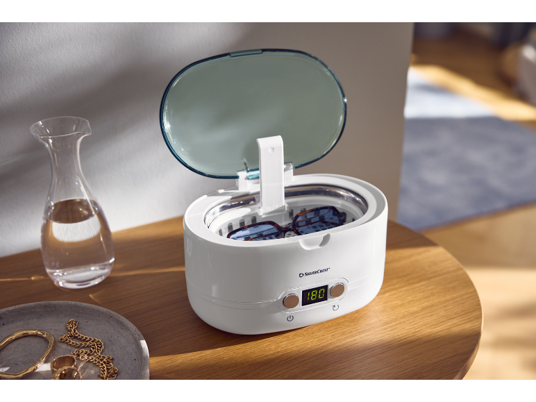 Ultraschallreiniger mit Brille auf einem Holztisch, neben einer Karaffe und Schmuck.