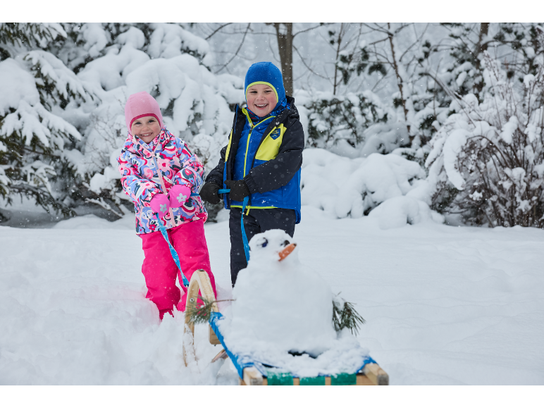 Zwei Kinder in Winterkleidung mit Schlitten und kleinem Schneemann in verschneiter Landschaft.