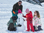 Eine Familie baut einen Schneemann im Schnee, die Kinder tragen rote und grüne Winterkleidung.