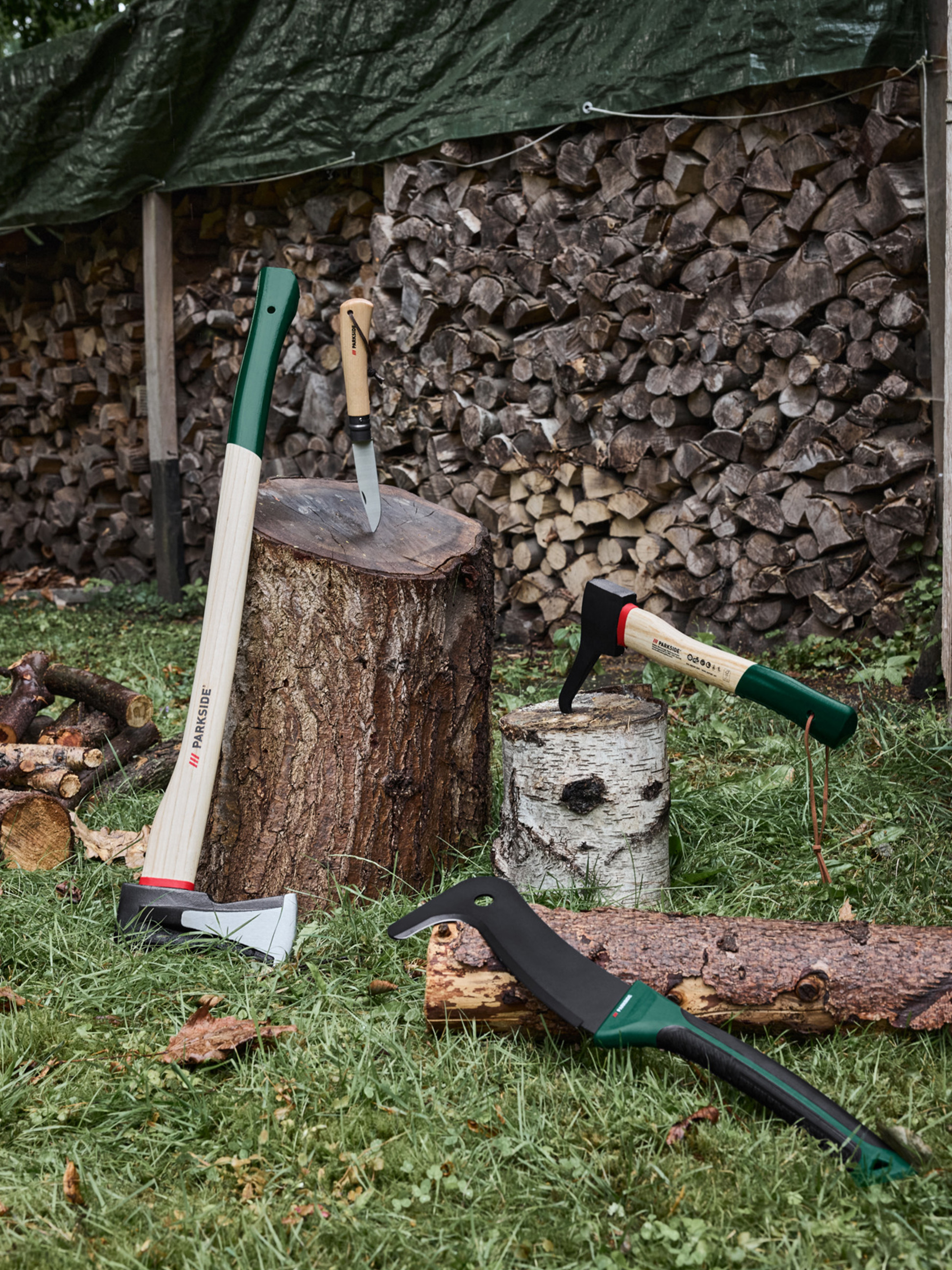 Äxte, ein Messer und eine Machete auf Gras mit gestapeltem Brennholz im Hintergrund.